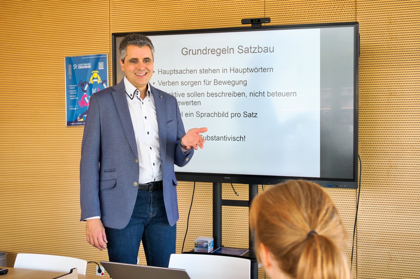 Daniel Völpel in weißem Hemd und blauem Sportsakko steht vor einem großen Bildschirm vor einer gelb-schwarz gerasterten Wand. Er spricht und gestikuliert in Richtung einer Zuhörerin, deren Kopf am unteren Bildrand zu erkennen ist.
