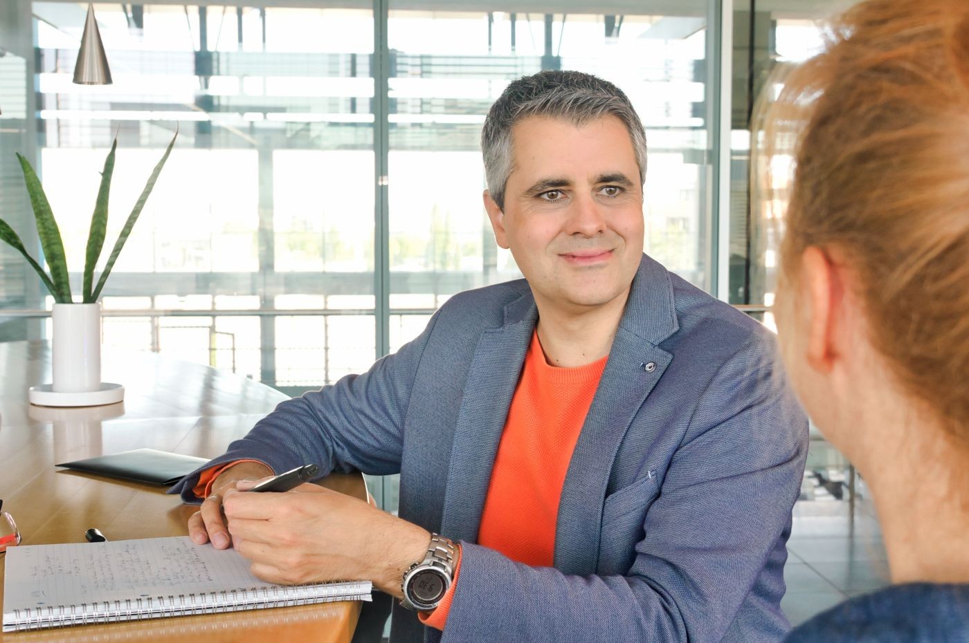 Über die Schulter einer Frau fotografiert, groß in der Bildmitte ein Mann, Daniel Völpel, gekleidet in orangenem Pullover und blauem Sportsakko. Beide sitzen an einem Tresen, er hat einen Block vor sich liegen. In der Linken hält er über dem Block den Stift und blickt die Frau an.