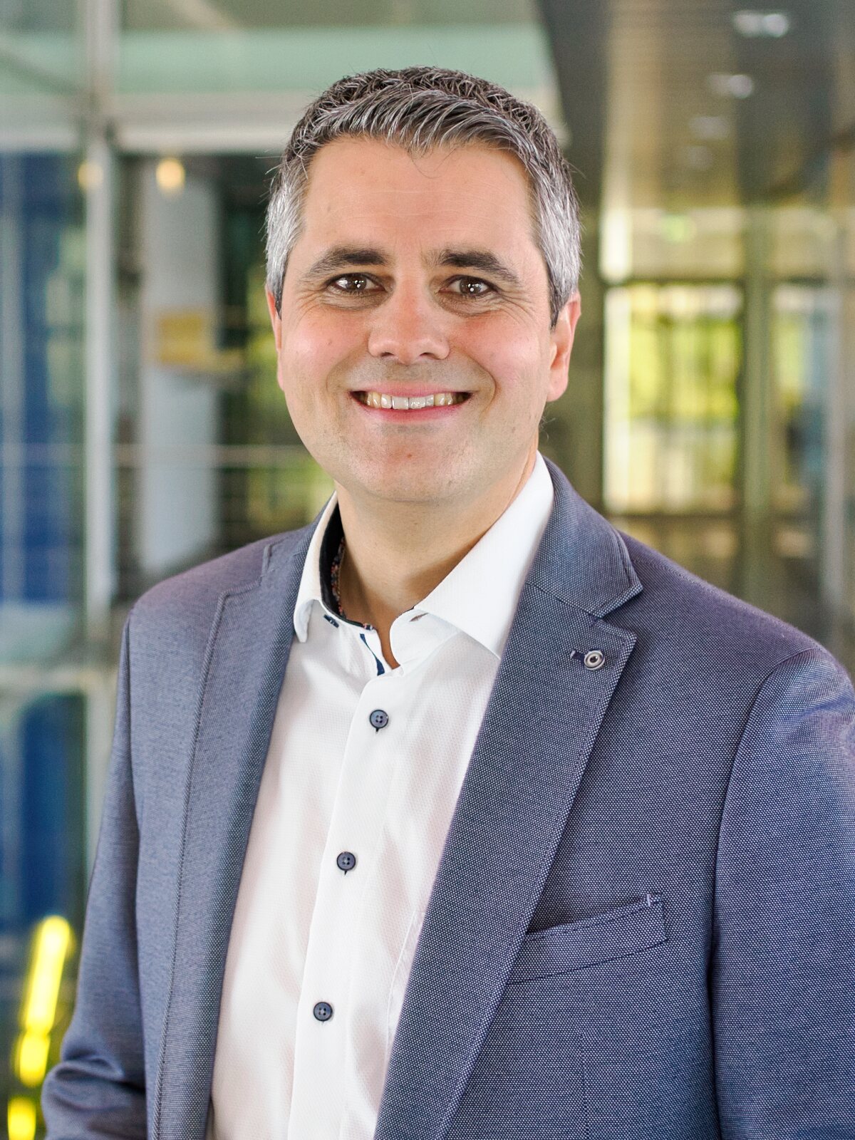 Daniel Völpel steht in einem unscharf erkennbaren Treppenhaus in Großaufnahme, blaues Sakko, weißes Hemd, und lächelt in die Kamera.